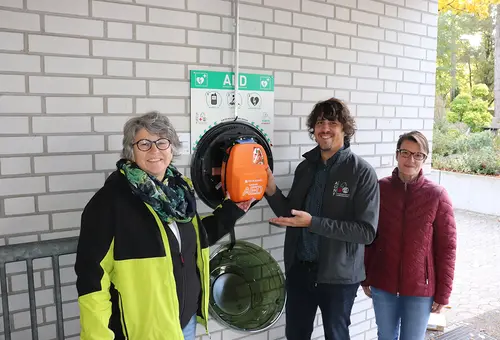 Zwei Frauen und ein Mann zeigen auf den neuen Wand-Defibrillator.