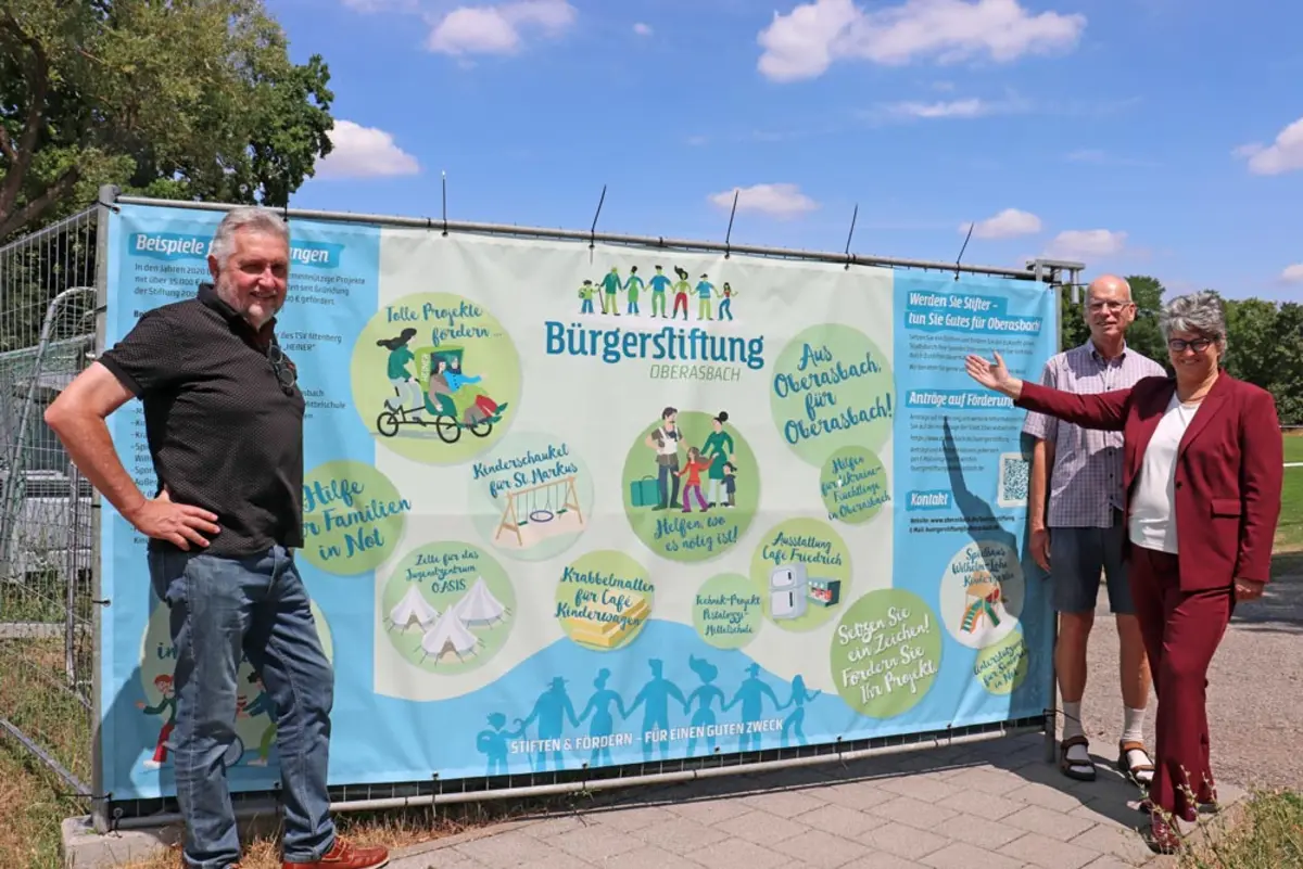 Zwei Männer und eine Frau vor einem Bauzaunbanner