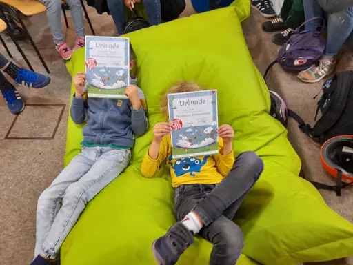 Zwei Kinder auf einem hellgrünen Sitzsack halten Bücher vors Gesicht