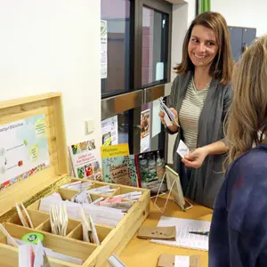 Eine junge Frau zeigt einem von hinten zu sehenden Mädchen einige Samentüten aus einer vor ihnen stehenden Holzkiste.