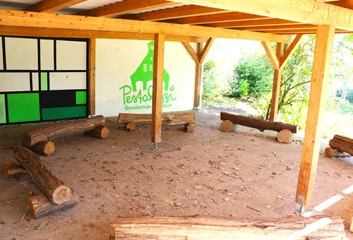 Blick in das Freiluftklassenzimmer, das nur an zwei Seiten von Wänden umgeben ist. Eine Holzkonstruktion hält das Flachdach darauf. Die Bänke sind Holzstämme mit rustikalem Touch.