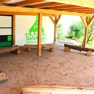 Blick in das Freiluftklassenzimmer, das nur an zwei Seiten von Wänden umgeben ist. Eine Holzkonstruktion hält das Flachdach darauf. Die Bänke sind Holzstämme mit rustikalem Touch.