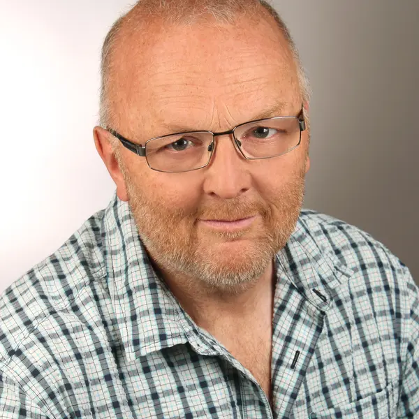 Porträt eines Mannes mit kurzen grauen Haaren, kurzem Vollbart und einem karierten Hemd. 