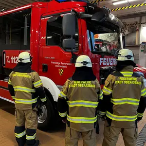 Drei Feuerwehrmänner in Einsatzkleidung mit Helm in der Rückenansicht vor dem Feuerwehrfahrzeug.