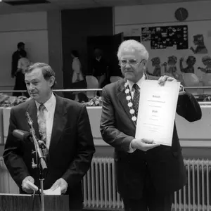 Schwarzweiß-Foto von Beckstein (links) und Güllering. Güllering hält die Urkunde zur Stadterhebung hoch. Vor Beckstein steht ein Mikrofon.