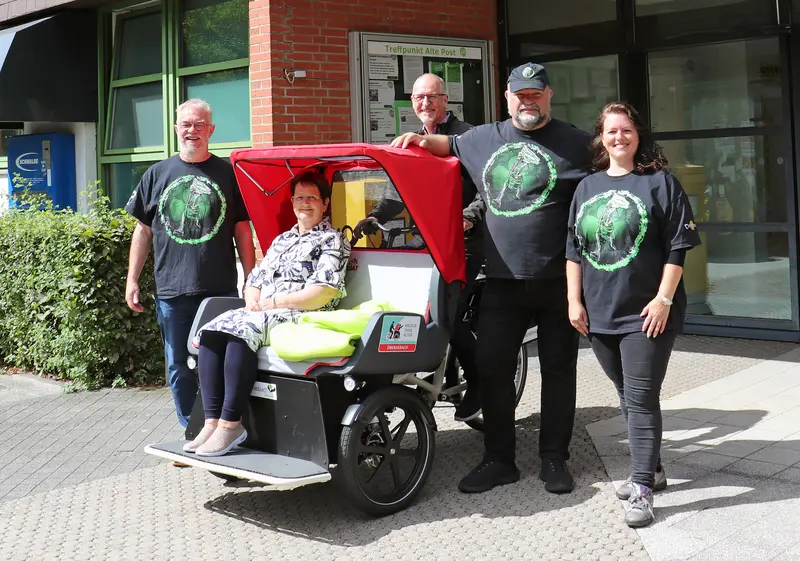eine Frau sitzt in einer Rikscha mit rotem Verdeck. Unm die Rikscha herum stehen drei Männer und eine Frau, die Rikscha-Pilotinnen. 