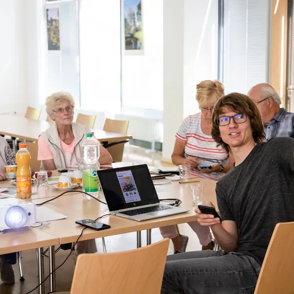 Ein Seminarraum. Vorne erklärt ein Junger Mann mit Laptop etwas. Im Hintegrund sieht man Senioren.
