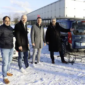 Drei Männer und eine Frau im Schnee neben einem Elektro-Auto, das an einer Ladesäule lädt. Im Hintergrund ein Gebäude. 