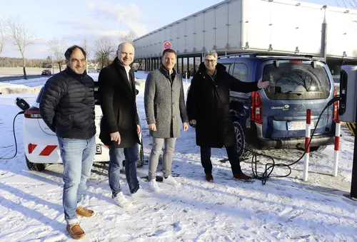 Drei Männer und eine Frau im Schnee neben einem Elektro-Auto, das an einer Ladesäule lädt. Im Hintergrund ein Gebäude.
