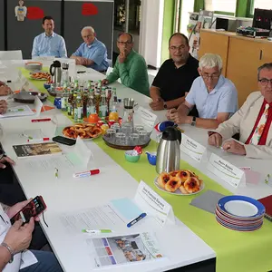 Arbeitsrunde: Am Tisch sitzen Männer und FRauen, die auf den nicht im Bild befindlichen Bildschirm schauen, über den die Videokonferenz nach Europa läuft.