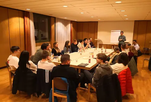 Im Hintergrund steht ein Moderator am Flip Chart. Im Vordergrund sitzen rund 20 Personen rund um einen großen Arbeitstisch.