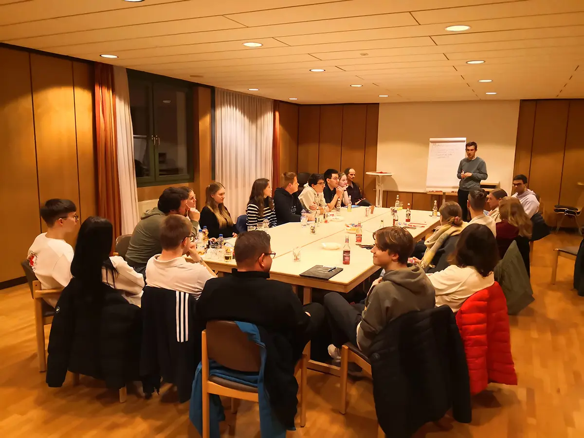 Im Hintergrund steht ein Moderator am Flip Chart. Im Vordergrund sitzen rund 20 Personen rund um einen großen Arbeitstisch.