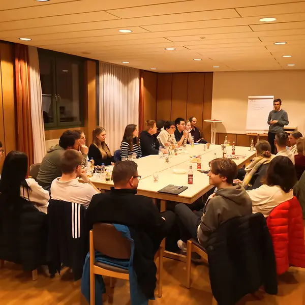 Im Hintergrund steht ein Moderator am Flip Chart. Im Vordergrund sitzen rund 20 Personen rund um einen großen Arbeitstisch.