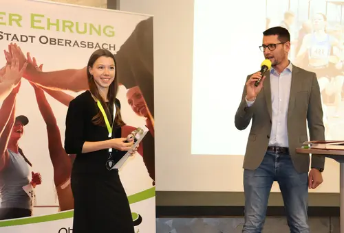 Eine junge Frau steht vor einer Plakatwand und ein Mann steht mit Mikrofon neben ihr.