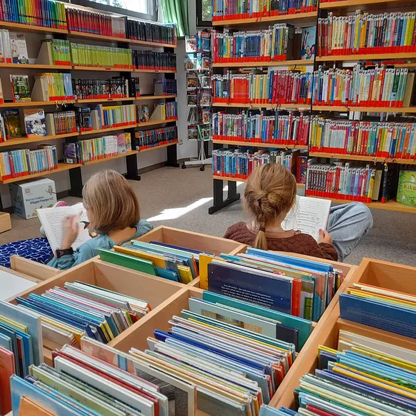 Zwei Mädchen sind mitten in der Bücherei und umgeben von Bücherregalen und dem Büchertrog in zwei Bücher vertieft.