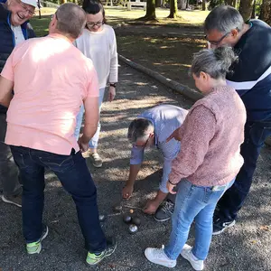 Fünf Personen stehen auf einer Boulebahn neben einem Mann, der auf dem Boden kniet und den Kugelabstand misst.