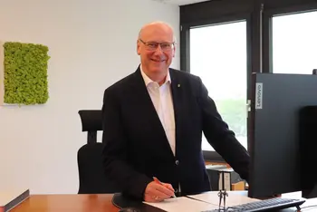 Mann mit weißem Hemd und Anzugjacke steht hinter einem Schreibtisch und lächelt freundlich. 