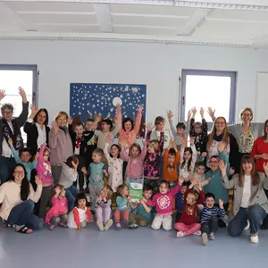 Fröhliche Kinder beim Gruppenbild, strecken die Hände nach oben, umrahmt von zehn erwachsenen Frauen.