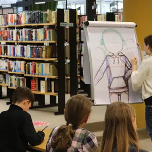 Workshop-Leiterin vor einem Flipchart, auf dem ein Rumpf und die Umrisse eines Kopfes zu sehen sind. Im Vordergrund die Rücken dreier Kinder, die am Kurs teilnehmen.
