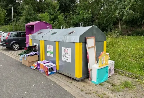 Blick auf einen Altglascontainer, vor dem Kartons, Kinderküchen, Flachbildschirme u.v.m. abgestellt wurden.