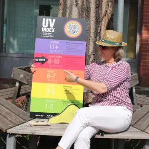 Frau mit Sonnenhut und -brille weist auf Tafel mit UV-Index