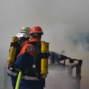 Zwei Jugendliche in Feuerwehr-Montur löschen einen Brand. Um sie herum Rauch- und Dampfschwaden.