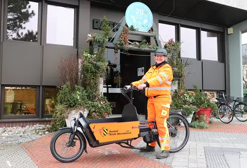 Frau in orangefarbener Arbeitsbekleidung auf dem Lastenrad vor dem Rathauseingang.