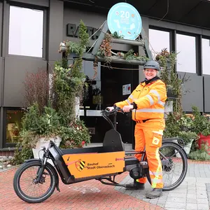 Frau in orangefarbener Arbeitsbekleidung auf dem Lastenrad vor dem Rathauseingang.