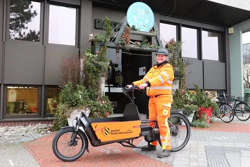 Frau in orangefarbener Arbeitsbekleidung auf dem Lastenrad vor dem Rathauseingang.