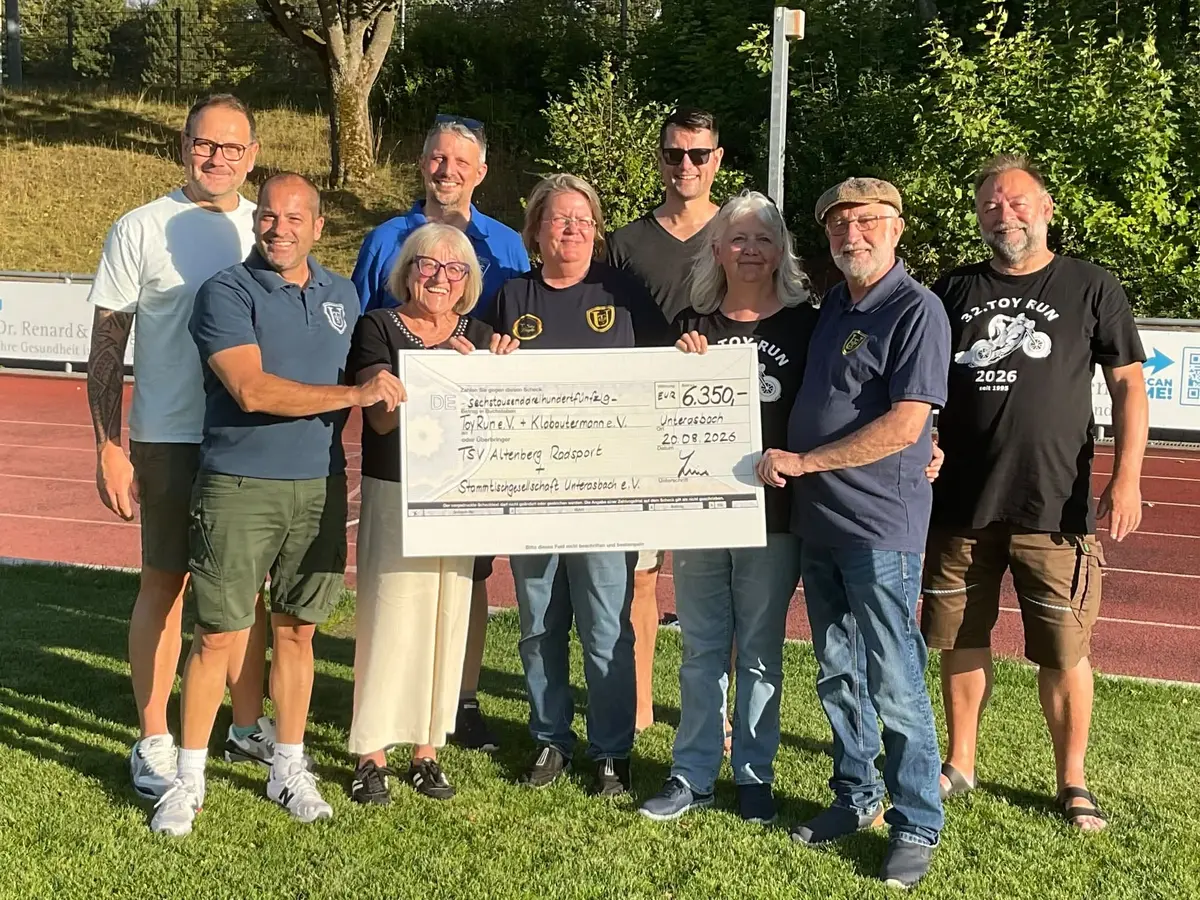 6 Männer und 3 Frauen halten auf einem Sportplatz einen großen, symbolischen Spendencheck über 6350 Euro. 