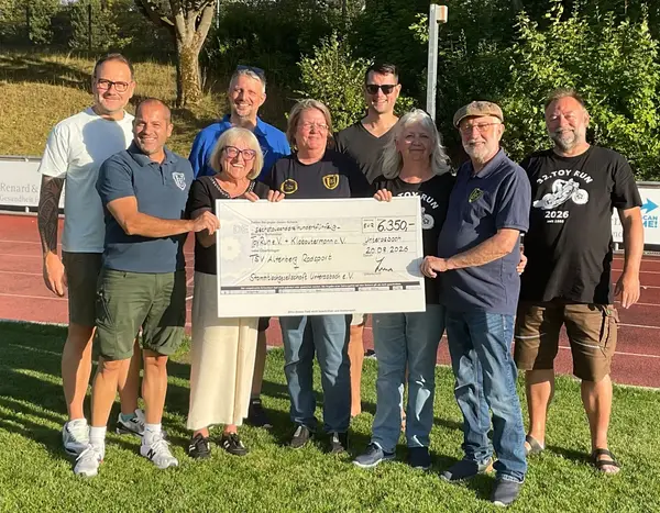 6 Männer und 3 Frauen halten auf einem Sportplatz einen großen, symbolischen Spendencheck über 6350 Euro. 