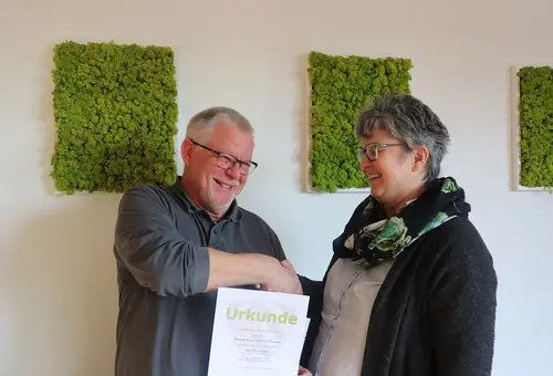 Eine Frau schüttelt einem Mann die Hand und überreicht eine Dankurkunde.