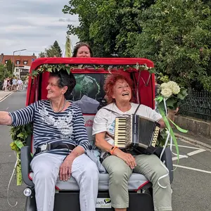 Zwei fröhliche ältere Damen, eine von ihnen mit Akkordeon, sitzen in einer Elektro-Fahrrad-Rikscha, die von einer Pilotin gefahren wird, und winken den Schaulustigen bei einem Festumzug zu.