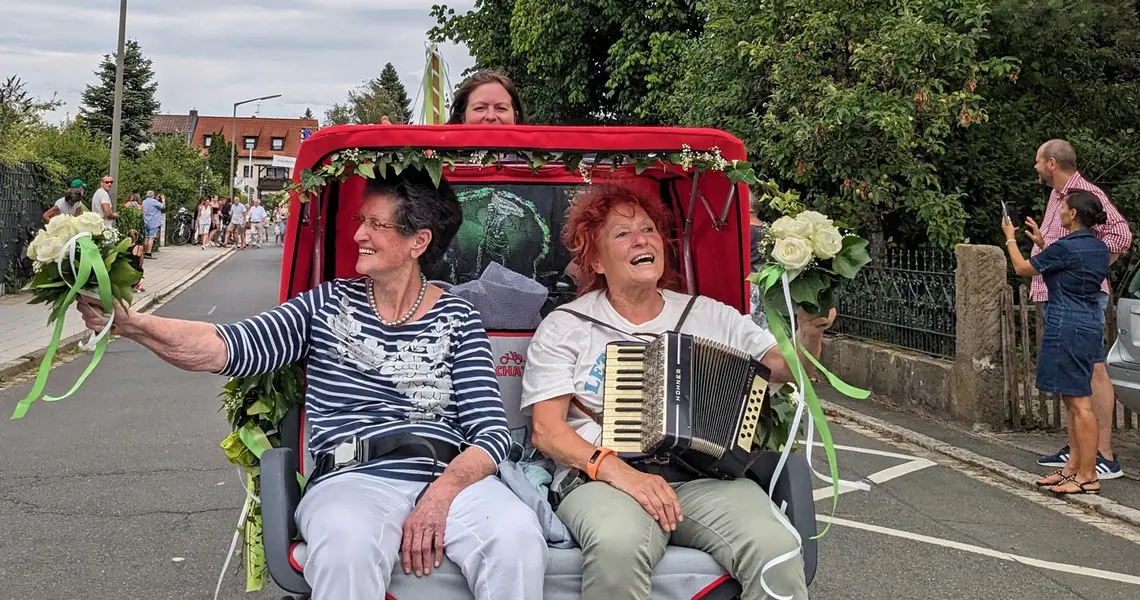 Zwei fröhliche ältere Damen, eine von ihnen mit Akkordeon, sitzen in einer Elektro-Fahrrad-Rikscha, die von einer Pilotin gefahren wird, und winken den Schaulustigen bei einem Festumzug zu.