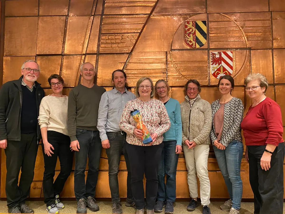 Neun Frauen und Männer vor einem Kupfer-Relief, die Dame in der Mitte hält ein Abschiedsgeschenk