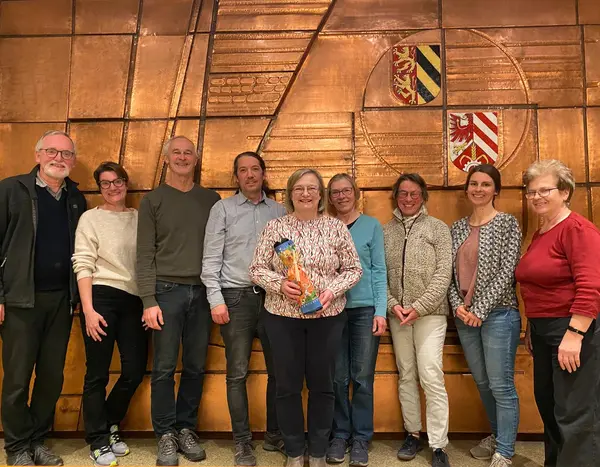 Neun Frauen und Männer vor einem Kupfer-Relief, die Dame in der Mitte hält ein Abschiedsgeschenk