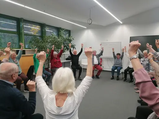 Eine Gruppe von Senioren sitzt mit erhobenen Armen auf Stühlen in einer Runde, vielen lachen.