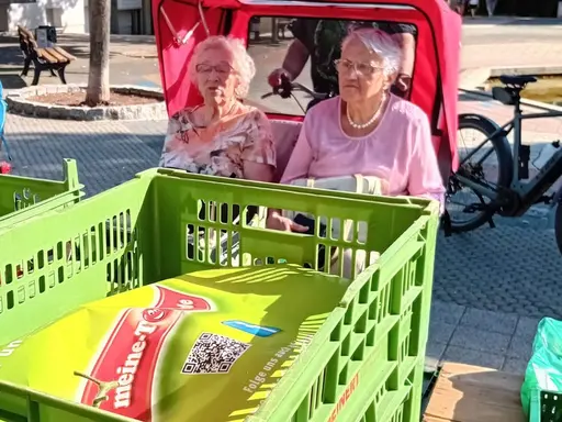 Zwei Seniorinnen sitzen in einer Fahrradrikscha und betrachten Gemüse auf dem Wochenmarkt 