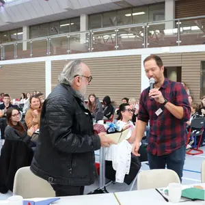 Ein älterer Mann hält einen Präsentkorb in der Hand. Ein jüngerer Mann gratuliert ihm. 