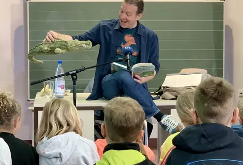 Autor Kröner sitzt auf dem Lehrerpult und unterhält sich mit seinem Stoffkrokodil. Davor Schuler:innen, die gebannt zuhören. 