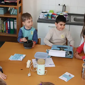 sechs KInder sitzen um einen Tisch herum, drei Kinder hantieren mit Mörser, Schreibmaschine und Kaffeemühle.