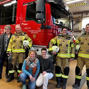 Drei Männer in Feuerwehreinsatzkleidung mit drei Frauen vor einen Einsatzfahrzeug