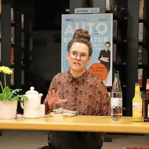 Katja Diehl an einem Tisch sitzend. Im Hintergrund ein Plakat zu ihrem Buch "Autokorrektur". 