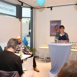 Frau an Stehstisch spricht zu Menschen, die um Tische sitzen