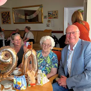 Bürgermeister, Jubilarin und Gäste. Auf dem Tisch im Vordergrund bilden zwei goldene Luftballons die Zahl 90. 