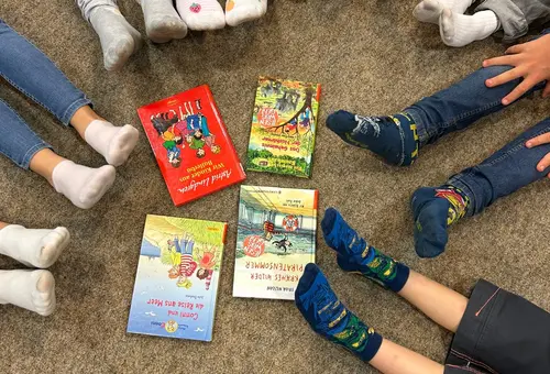 Vier Bücher in der Mitte des Bildes und darum herum die Beine und Füße der Teilnehmer:innen.