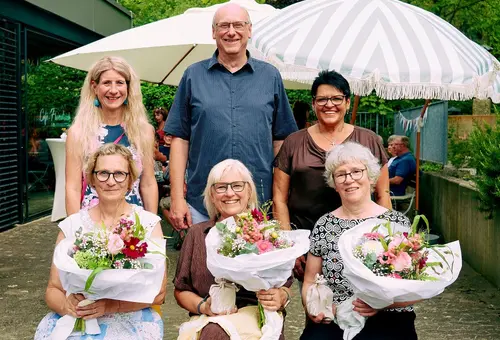 Drei Damen sitzend mit Blumenstrauß, dahinter ein Mann und zwei Frauen