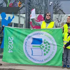 Eine Frau zeigt eine Urkunde und zwei Kinder halteneine große grüne Flagge. Eine weitere Person steht daneben.