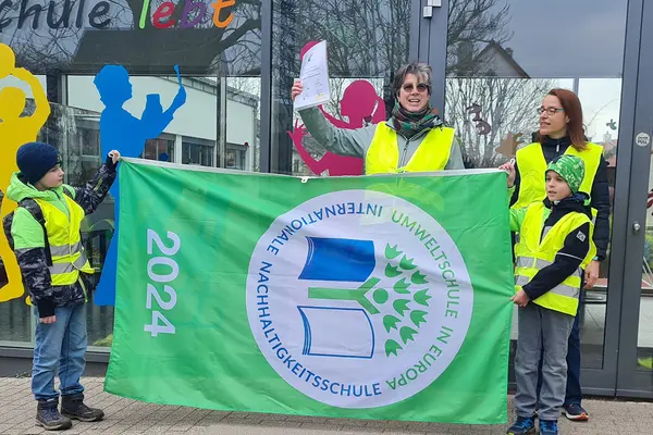 Eine Frau zeigt eine Urkunde und zwei Kinder halteneine große grüne Flagge. Eine weitere Person steht daneben.