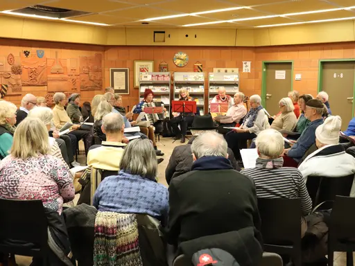 Sängerinnen und Sänger sitzen im Foyer des Rathauses im Kreis. In der Mitte Musikerinnen mit Geige, Akkordeon und Gitarre. 